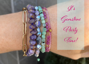 A stack of bracelets- a heavy gold chain by Maria Beaulieu, a purple gemstone bracelet by Joseph Brooks, a purple pearl and opal bracelet, a faceted square emerald beaded bracelet and two delicate ruby bracelets