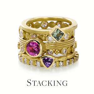 A stack of 18k yellow gold bands by jewelry designer Barbara Heinrich; from top to bottom: a smooth, square band with a bezel set green tourmaline, an alternating round gold bead and round diamond band, a grooved band with a round, faceted, bezel set hot pink tourmaline, a pillow band with a trillium, bezel set purple sapphire and a diamond gear band