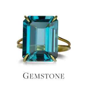 A 22k yellow gold ring by jewelry designer Rosanne Pugliese featuring a central prong-set, emerald-cut London Blue Topaz stone on a split shank. The piece sits on a white background above the word "Gemstone," and links to all rings in the "Gemstone" category available on the website. 