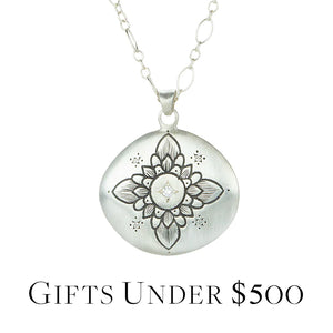 A sterling silver necklace by the jewelry designer Adel Chefridi featuring a chain with a round disc pendant with a flower engraved in black and accent with five bead set white diamonds. Link is to the "Gifts Under $500" collection