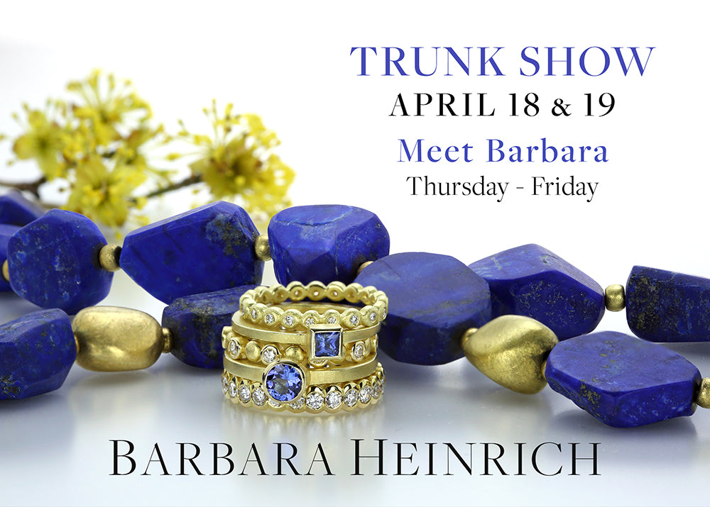 A large, dark blue, lapis nugget necklace with interspersed 18k yellow gold beads with a stack of yellow gold and diamond rings in the foreground, one with a round, faceted blue sapphire and one with a square, bezel set blue sapphire