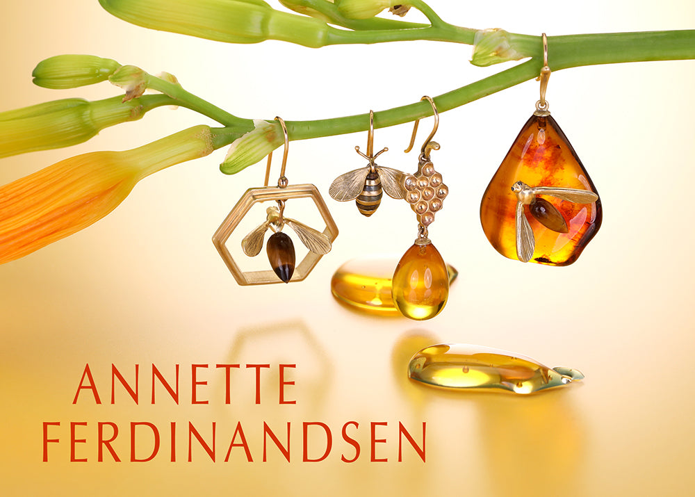 Three amber and gold jewelry pieces hanging from a green flower stem against a warm cream background, with the text 'ANNETTE FERDINANDSEN' in red at the bottom left.