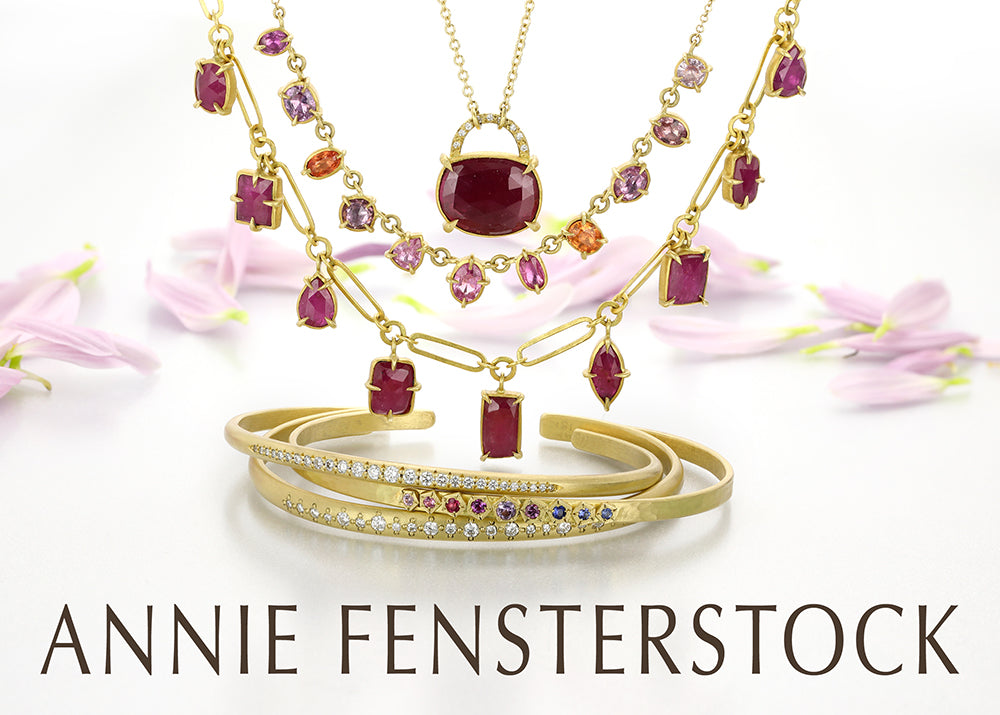 A stack of three diamond and sapphire bangles with three ruby and sapphire necklaces and pink petals in the background. Handcrafted by jewelry designer Annie Fensterstock