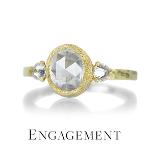 An 18k yellow gold engagement ring by jewelry designer Todd Pownell with a central rose-cut diamond set in a textured bezel and flanked by two prong-set inverted white diamonds on a textured band. The ring sits on a white background with a faded drop shadow above the text, "Engagement," which links to all rings which fall under the "Engagement" category currently on the website.