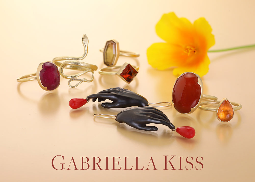 A styled product shot of several gold rings with red, black, and amber gemstones, arranged on a warm beige background with a yellow flower and the text 'GABRIELLA KISS' at the bottom.