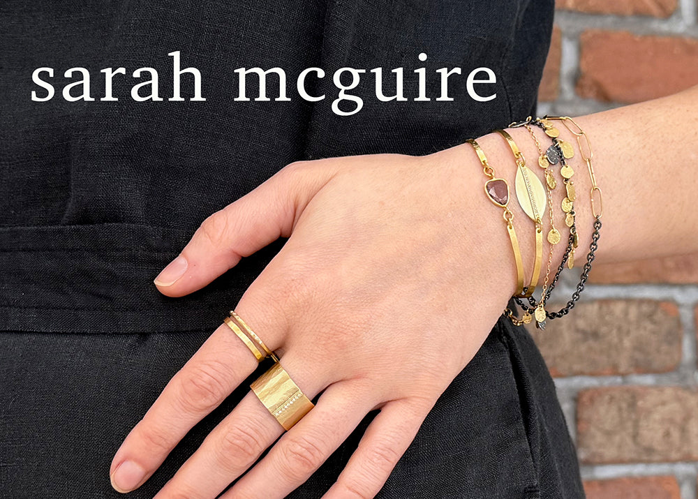 A close-up of a person's hand and wrist wearing multiple gold rings and bracelets, including a leaf-shaped charm bracelet, over a black top, with a brick wall in the background and the text 'sarah mcguire' at the top.