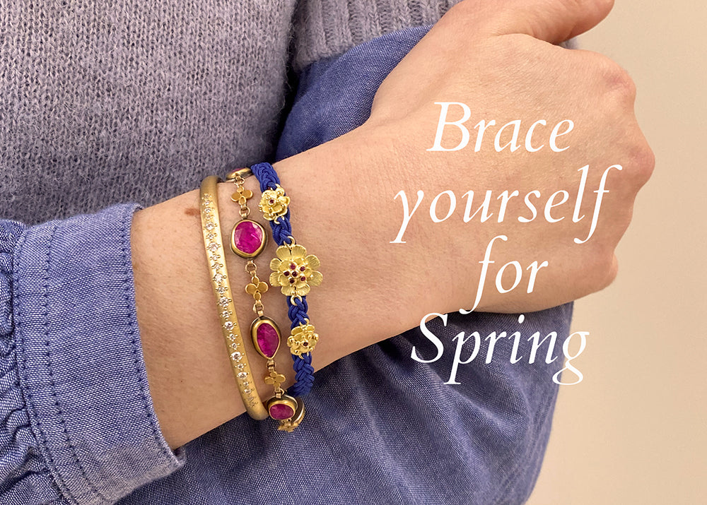 A model wearing a purple sweater and a denim shirt with three bracelets on: an 18k yellow gold and diamond bangle by jewelry designer Annie Fensterstock, a gold and faceted ruby bracelet by jewelry designer Ananda Khalsa and a blue woven cord bracelet with three flower elements by jewelry designer Lene Vibe. 